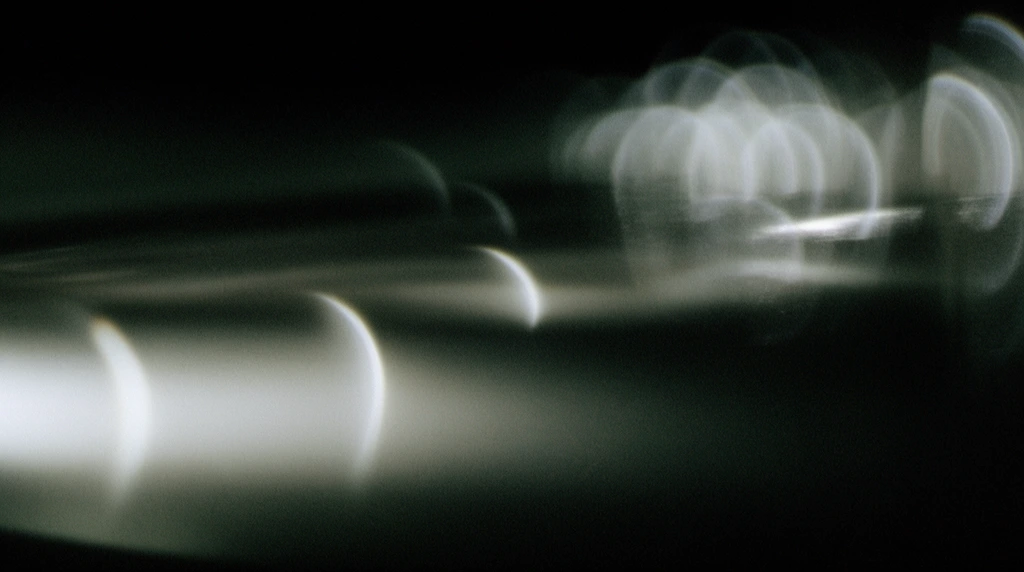 Abstract out-of-focus light streaks sweep horizontally across a pitch-black background. Soft, glowing white gradients blend with distinct crescent-shaped bokeh highlights scattered along the dark expanse. A hazy cluster of interconnected luminous arcs dominates the upper right quadrant. The composition simulates macro photography of light reflecting off a smooth, glossy surface, featuring heavy cinematic film grain, dark greenish-grey undertones, and anamorphic lens aberrations that create a moody, highly textured atmospheric study of diffuse illumination.