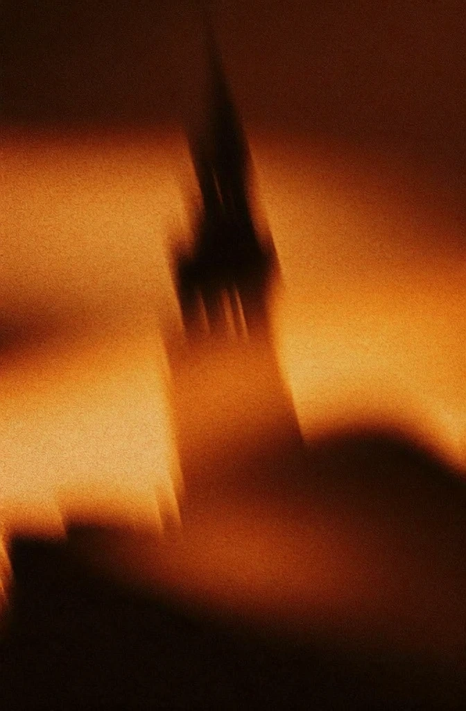oblique silhouette of a monolithic bell tower and spire rising above a dense urban cluster, rendered in a high-chroma amber and burnt sienna monochromatic palette, saturated with heavy stochastic noise and gritty film grain, extreme intentional camera movement creating soft motion blur, out-of-focus ethereal atmosphere, minimalist composition where the architecture is an amorphous dark mass against a glowing hazy background, high-contrast noir aesthetic, deeply textured.