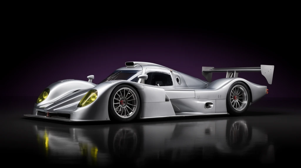 silver prototype endurance racing car, extreme low profile, sweeping aerodynamic body, flared fenders, multi-spoke wheels, large rear wing, roof scoop, yellow-tinted headlights, premium automotive studio photography, exact side profile, pitch black background, subtle purple gradient, glossy reflective floor, high contrast lighting