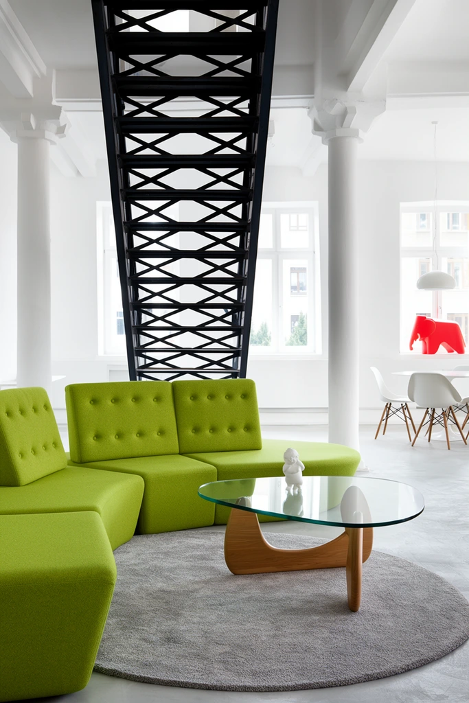 modern minimalist interior bathed in bright daylight, lime green modular sofa with button-tufted backrest on a circular grey area rug, organic-shaped glass coffee table with curved wooden base, small white figurine on the table, prominent industrial black metal open-tread staircase with cross-bracing, white walls and thick structural columns, background dining setup with white table and molded plastic chairs, large windows, red plastic elephant sculpture on sill, clean architectural photography