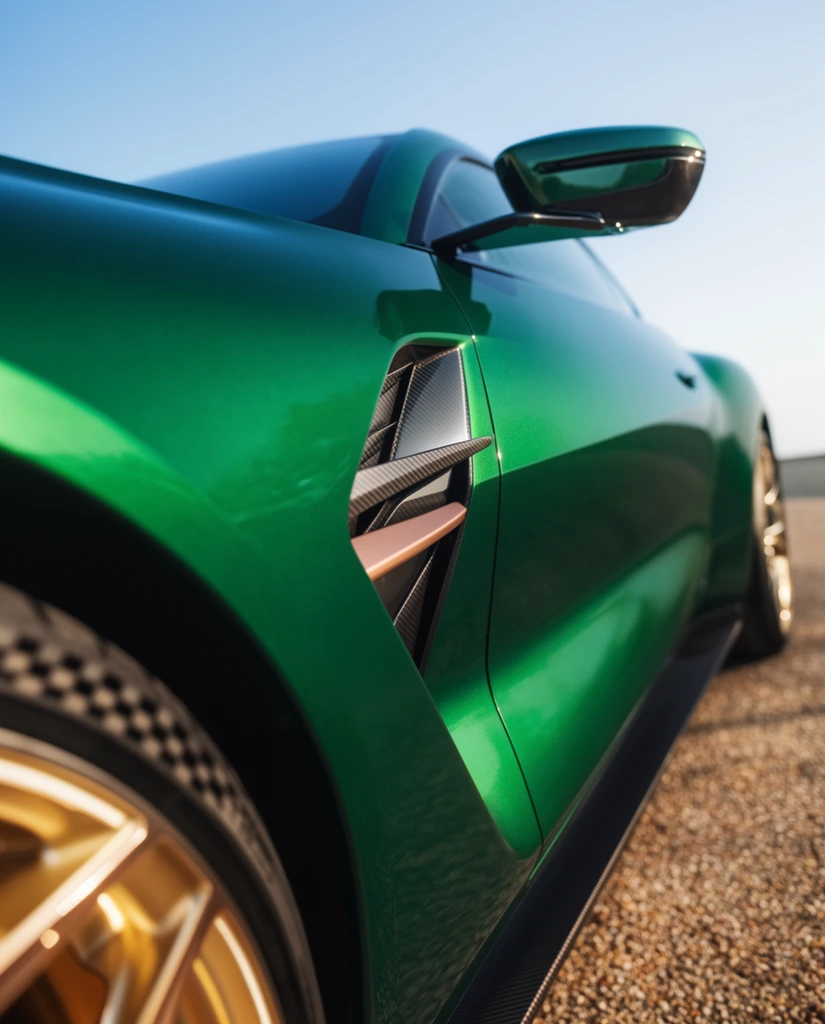 A dramatic, close-up view captures the aerodynamic side profile of a metallic emerald green luxury coupe under clear daylight. The extremely low-angle composition focuses on the front fender, looking down the deeply sculpted door panel to emphasize the vehicle's sweeping, aggressive lines. A recessed side air intake featuring sharp, geometric carbon-fiber louvers and a brushed bronze trim piece sits sharply in focus on the quarter panel. A sharply angular side mirror mounted on a slender stalk extends outward, catching the bright sunlight. In the extreme foreground, a partially visible tire with a checkered tread pattern wraps around a gleaming gold multi-spoke alloy wheel. The background fades into a smooth, shallow depth of field, revealing a gradient light blue sky and a warm, textured gravel surface. Brilliant natural lighting creates intense specular highlights across the glossy metallic paint.