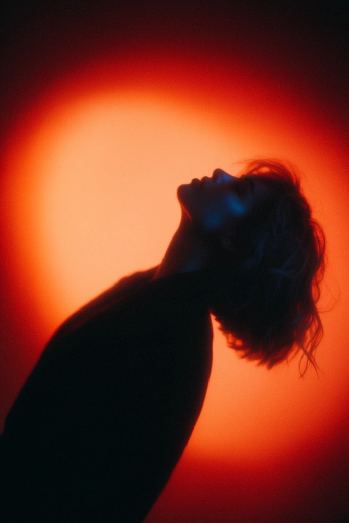 A cinematic, high-contrast portrait featuring the dark silhouette of a figure against a vibrant glowing background. The person is captured from the torso up, leaning backward with an arched spine and head tilted upwards in a striking profile. They wear a dark garment. Their short, wavy hair is softly textured with loose strands catching the light. The background is an intense, diffused circular light source grading from bright orange in the center to deep red at the edges, creating a dramatic halo effect. A subtle, cool blue rim light highlights the edge of the subject's facial profile, contrasting sharply with the warm backdrop. The image possesses a moody, atmospheric quality characterized by visible film grain and a slightly soft focus.