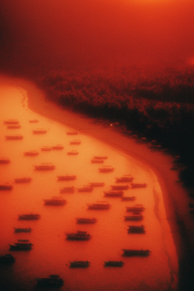 An aerial view of a coastline reveals numerous small boats anchored in the water near a beach and forested land, drenched in a heavily grained, intensely glowing monochromatic palette of fiery reds and oranges. From a high vantage point, dozens of scattered boats rest on the water's surface, adjacent to a smooth coastal strip that meets a dense, textured forest. The boats, shoreline, and trees appear as deep, heavily underexposed, near-black silhouettes contrasting starkly against the radiant, luminous water and hazy environment. Saturated gradients of burnt amber, deep crimson, and bright tangerine flood the entire scene. A thick atmospheric haze softens all distinct edges, producing a dreamy, diffused halation around the darker shapes. The visual execution mimics vintage analog film, characterized by a prominent, coarse film grain, a soft optical focus, and a lo-fi ethereal aesthetic that emphasizes stark geometric silhouettes and overwhelming, glowing warmth over fine detail.