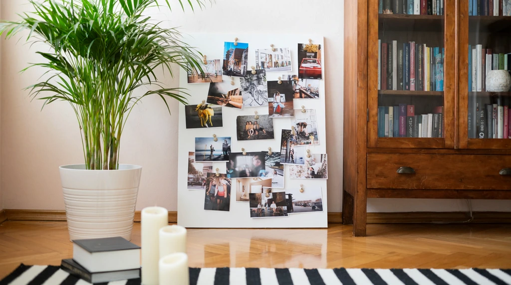 A bright interior featuring a large white mood board layered with pinned photographs leaning against a pale wall. Beside it sits a green palm in a white planter and a glass-fronted wooden bookcase. The foreground reveals three white pillar candles and a thick book near a black-and-white striped rug on a glossy hardwood floor, all bathed in soft natural daylight.