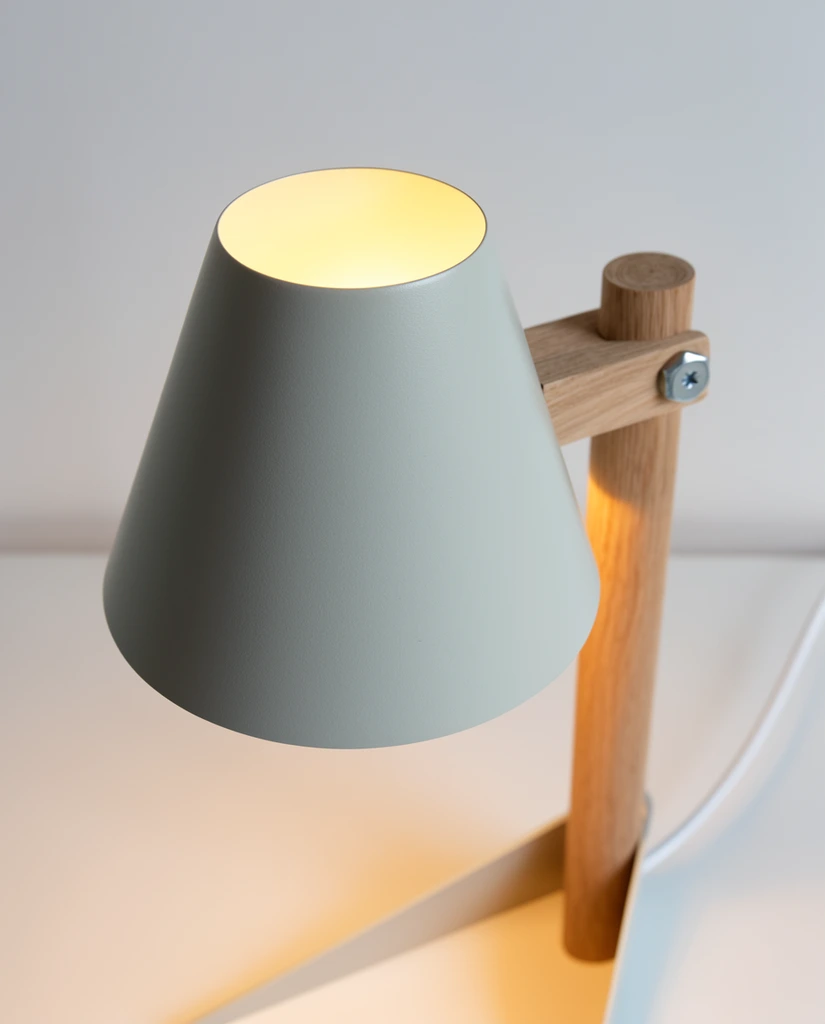 minimalist table lamp, matte pale grey powder-coated truncated cone shade, warm yellow light glowing from top circular opening and casting downward, natural light wood side arm, visible metal hex screw, flat angled metal base, white power cord, smooth white surface, plain off-white background, subtle shadow gradient, shallow depth of field, high-end Scandinavian product photography, close-up high angle shot