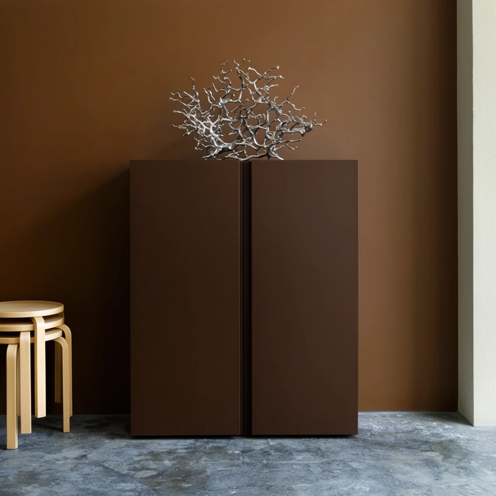 A minimalist interior featuring a dark brown two-door cabinet centered against a matching matte brown wall. The cabinet has clean geometric lines and rests on a mottled gray concrete floor. A complex, branching metallic silver sculpture sits atop the cabinet. On the far left, three light wooden round stools with curved legs are stacked together. A sliver of an off-white wall borders the right edge. Soft natural lighting creates subtle shadows, highlighting the strong color block contrast between the warm earthy tones and the cool textured concrete.