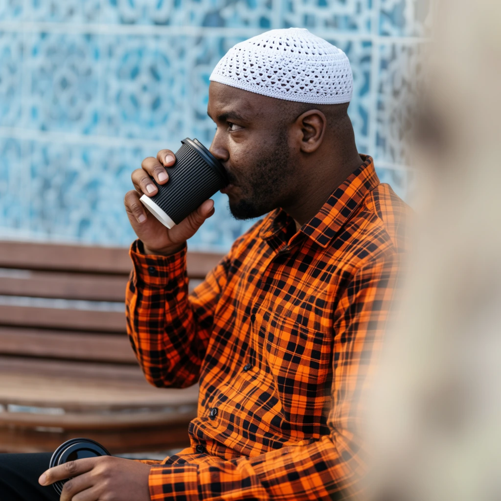 Black man in profile drinking from a black ribbed cup, wearing a white crocheted kufi and vibrant orange and black plaid shirt, hand resting on lap holding a black lid, seated near a wooden bench, soft bokeh background with blue geometric wall tiles, out of focus foreground element, shallow depth of field, natural daylight