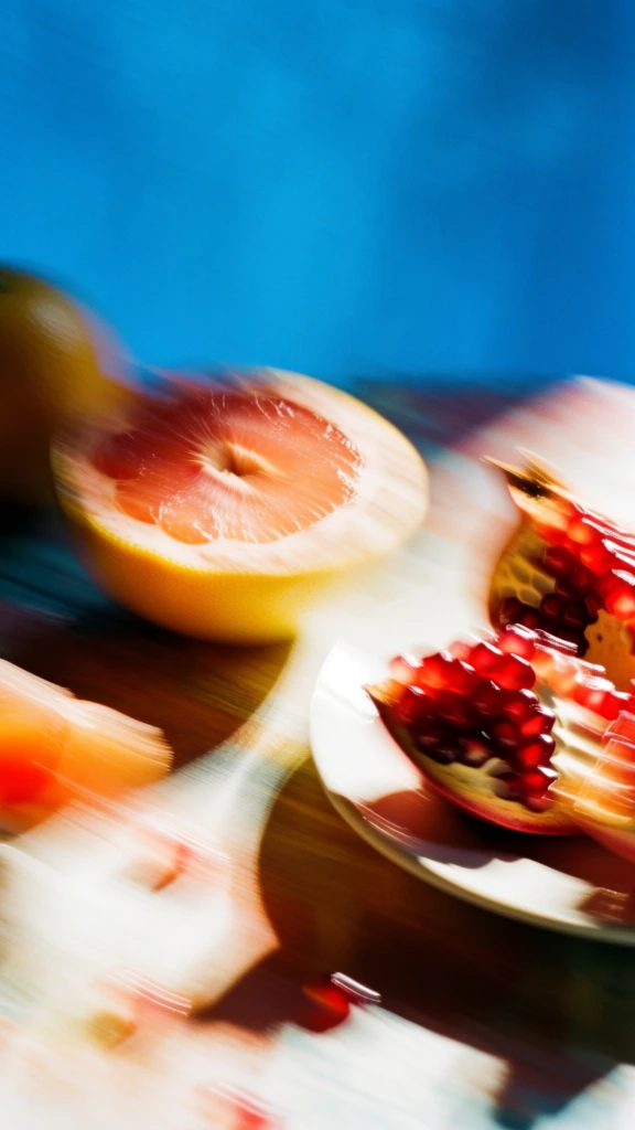 extreme motion blur with intentional camera movement, a grapefruit sliced in half and ruby pomegranate segments on a table, vibrant cerulean blue backdrop, high contrast directional lighting, saturated warm citrus tones versus cool background, analog film grain, dynamic diagonal sweeping streaks, soft abstract focus, cinematic high-key highlights, deep shadows, 35mm aesthetic, experimental still life, impressionistic texture, rich color saturation.