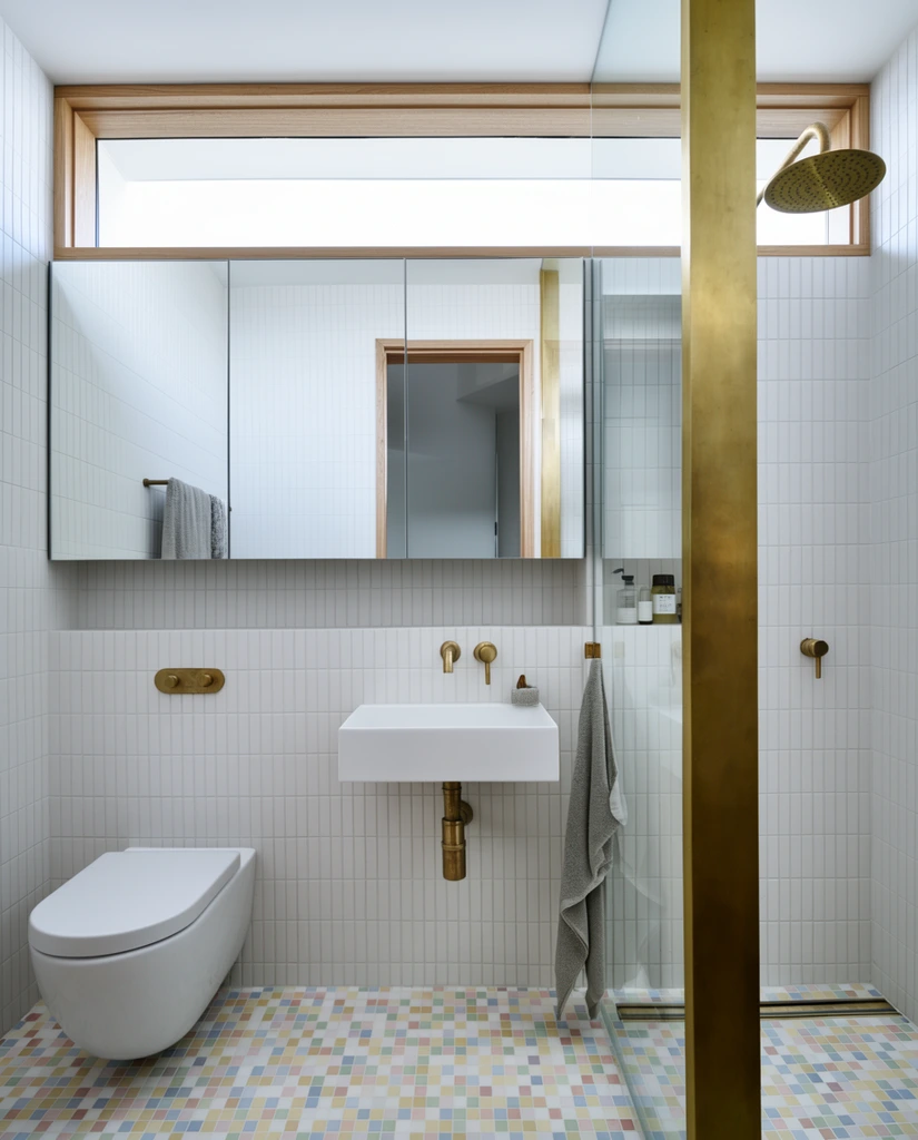 modern minimalist bathroom interior, wall-mounted white ceramic toilet, rectangular white wall-hung sink, exposed brushed brass plumbing and fixtures, vertical white wall tiles, multi-colored pastel square mosaic floor tiles, wide mirrored cabinet reflecting a doorway, narrow transom window with bright natural light, open shower area framed by a thick brass pillar, circular brass rain shower head, wooden trim, hanging grey towel, architectural photography, bright even lighting, crisp geometric lines