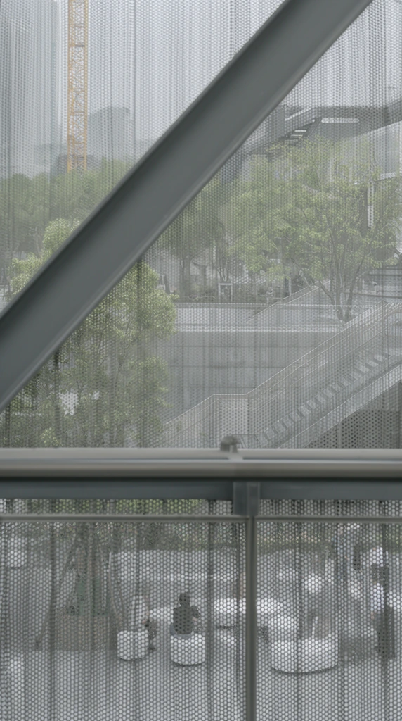 layered architectural view through a perforated metal screen, thick diagonal gray structural beam and horizontal railing, semi-transparent metallic mesh with dense circular holes creating a moiré pattern, lower section showing small figures near white cylindrical seating, upper background revealing a hazy urban plaza with green trees, modern staircases, distant yellow construction crane, muted gray and soft green color palette, diffused daylight, textural contrast, architectural photography