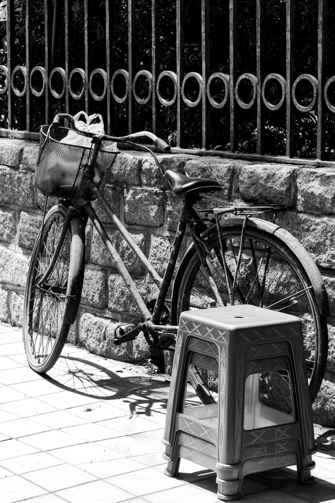 A well-worn bicycle leans against a textured stone wall beside a stack of plastic stools in a high-contrast black and white street photograph. The vintage bicycle features curved drop handlebars, a rear luggage rack, and a front wire basket holding a crumpled plastic bag, casting stark, intricate shadows onto the bright tiled pavement. Beside the rear wheel sits a stack of three square plastic stools with geometric X-patterned sides. The backdrop consists of rough-hewn stone blocks topped with a wrought-iron fence decorated with repetitive circular ring motifs, revealing dark foliage through vertical metal bars. The monochromatic composition emphasizes coarse textures and structural geometry, illuminated by harsh, direct sunlight that creates deep shadows and brilliant highlights, evoking a timeless urban documentary aesthetic.