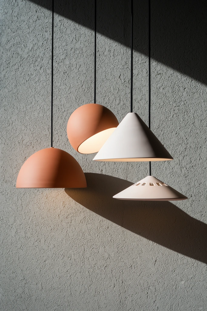 Four modern matte ceramic pendant lights hang at staggered heights against a heavily textured gray plaster wall. The smooth shades feature terracotta and soft white tones across diverse geometric forms: a classic dome, an angled spherical shell, a flared cone, and a shallow cone with small rectangular cutouts. Thin black cords suspend each warm-glowing fixture. Intense directional sunlight cuts a sharp diagonal line across the upper frame, casting stark, elongated shadows of the lamps onto the rough wall. The minimalist architectural composition highlights the contrast between the pristine curved silhouettes and the stippled background under striking, high-contrast lighting.