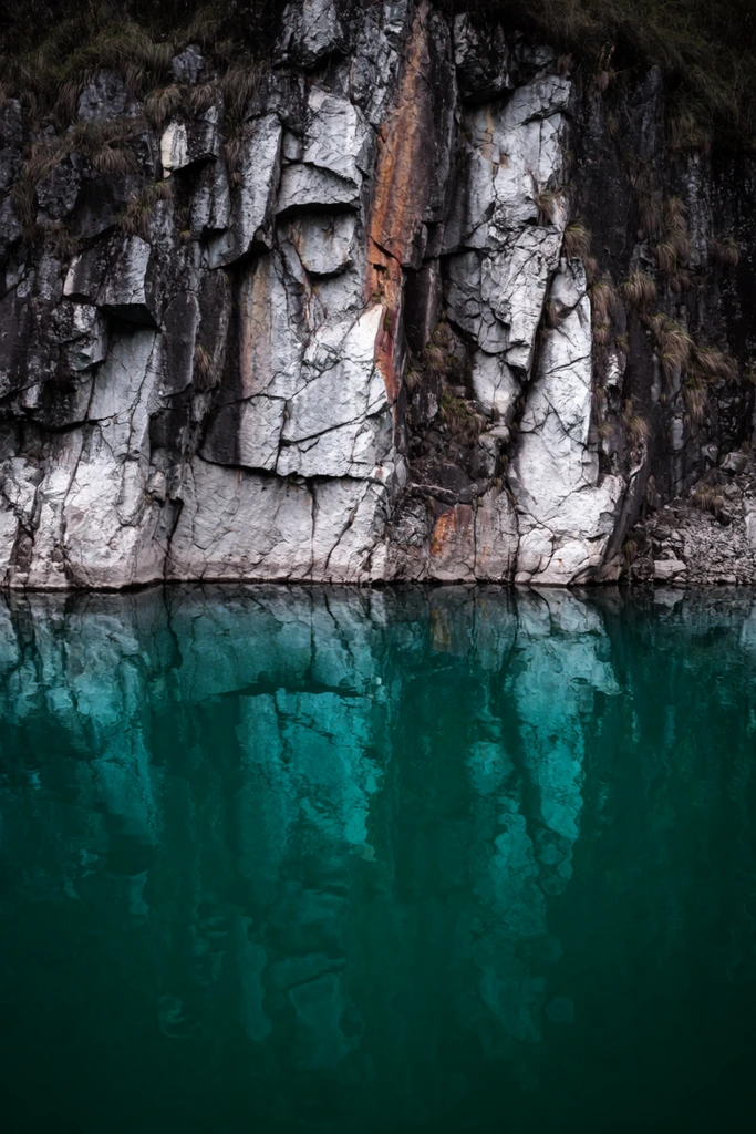 A sheer, rugged rock cliff face drops directly into a vibrant teal body of water. Deeply textured gray and white stone with vertical fissures, rust-colored stains, and tufts of dry grass fills the upper half. Below, calm emerald-green water creates a dark, wavy reflection of the imposing cliff. The stark horizontal split framing emphasizes the striking contrast between harsh jagged stone and smooth liquid in a moody, high-resolution natural landscape photograph.