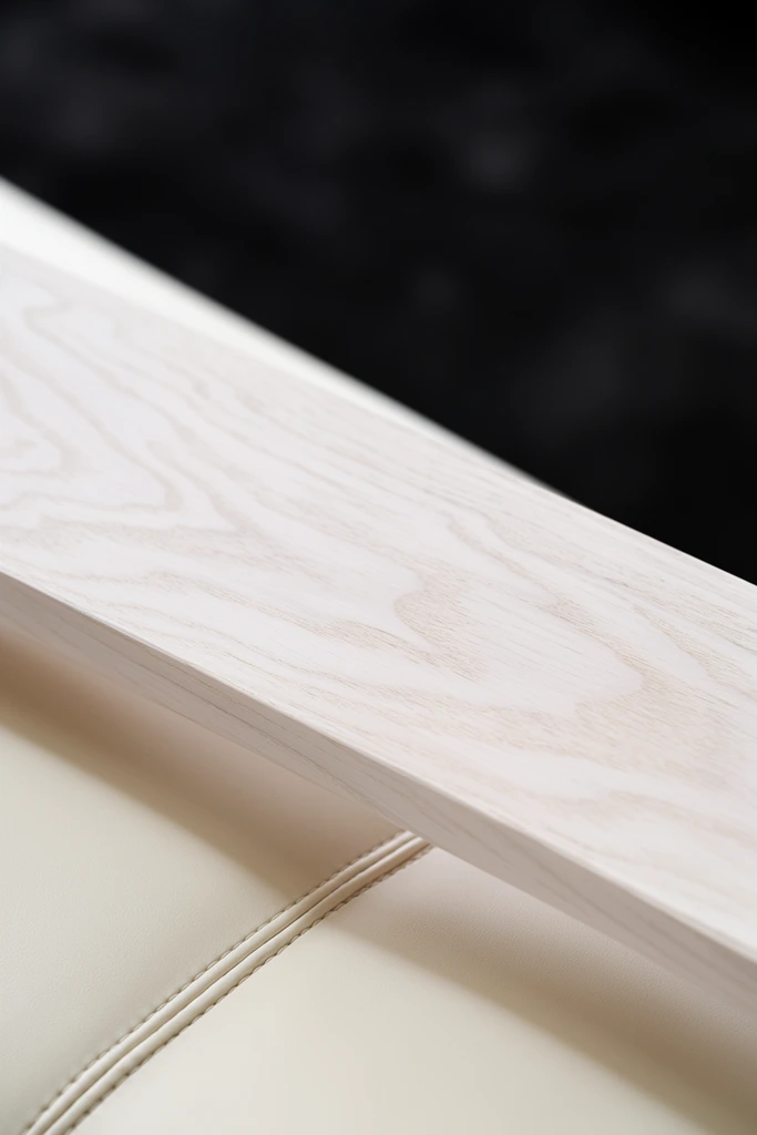 A minimalist macro photograph of an interior design detail. A pale, bleached wooden plank with delicate sweeping grain patterns extends diagonally across the frame. Directly below the wood lies a smooth, cream-colored upholstered surface detailed with an intersecting double-stitched seam. A soft, diffused shadow falls from the wooden edge onto the gentle curve of the upholstery. The upper portion of the composition drops off into a heavily blurred, deep charcoal black background. The shallow depth of field keeps the central wood grain in sharp focus while entirely softening the surrounding elements, emphasizing the tactile contrast between the organic wood, the stitched leather-like material, and the stark dark void.