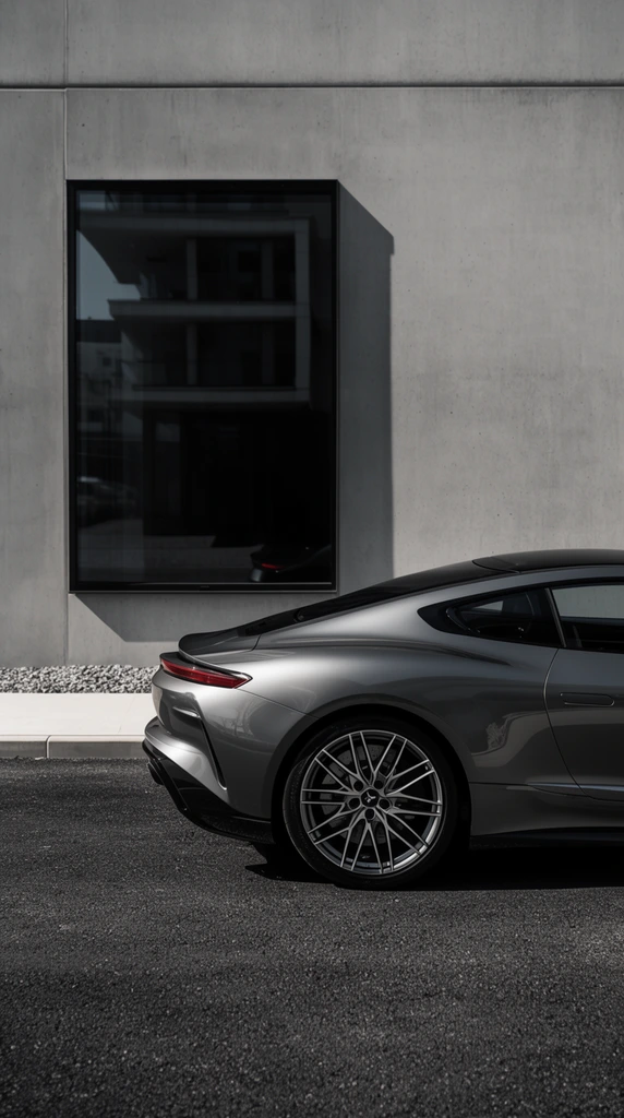 The rear profile of a metallic gray sports car rests on dark asphalt before a minimalist concrete building. The sleek vehicle features aerodynamic curves, a dark roofline, thin red taillights, and intricate multi-spoke alloy wheels. A light curb and gravel strip border a stark wall with a deeply tinted rectangular window reflecting an adjacent modern structure. Strong sunlight casts sharp shadows across the pavement and smooth facade, emphasizing the glossy automotive finish in a high-contrast, desaturated composition.