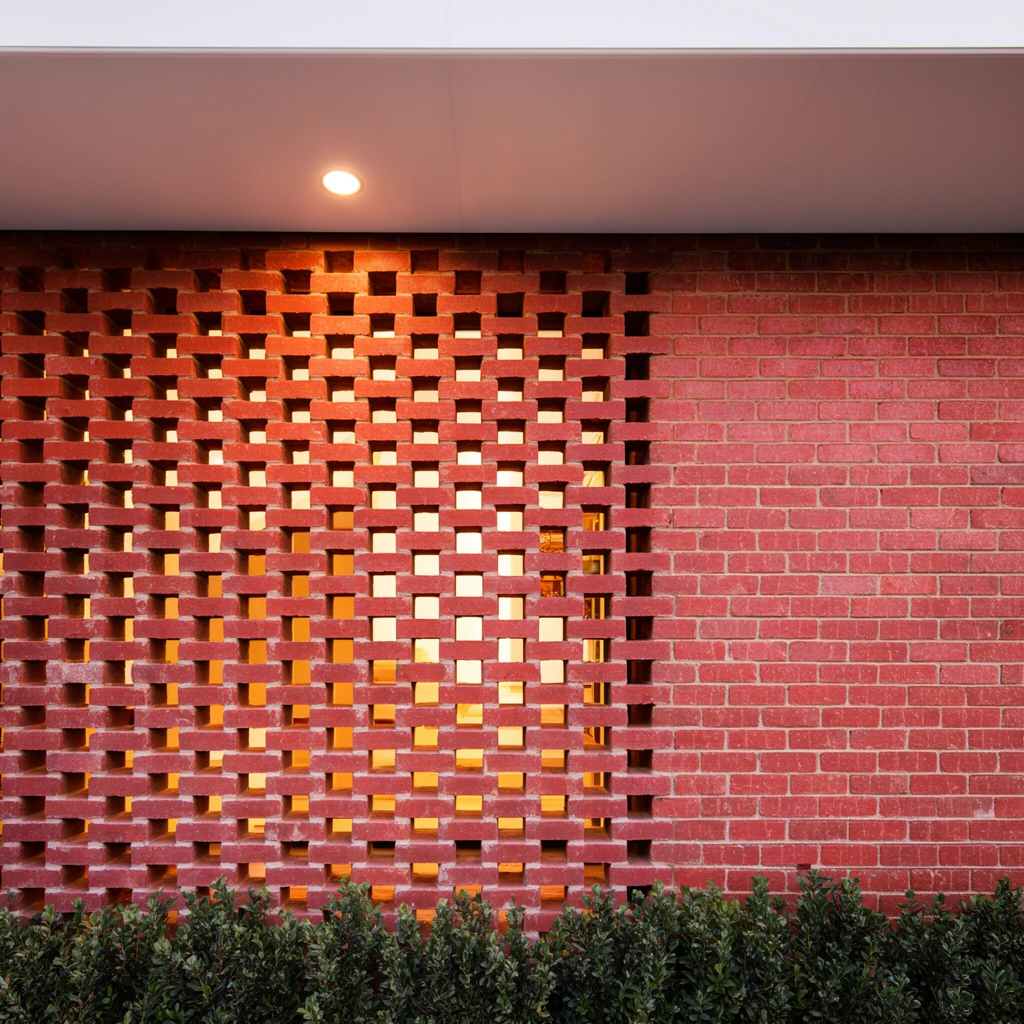 perforated red brick wall screen transitioning to solid masonry, warm interior light glowing through rectangular gaps, white overhang with single circular downlight, low green shrubs at base, architectural photography, straight-on elevation