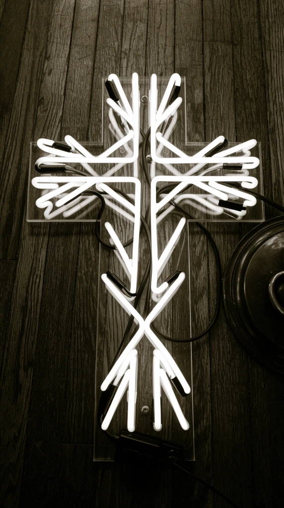 overhead perspective of an abstract cross-shaped white neon sculpture lying on a dark wooden plank floor, glowing glass tubes forming intersecting geometric branches, transparent acrylic backing board, thick black electrical cables weaving between the luminous tubes, small black power adapter at the base, edge of a dark circular metallic object on the right, dark stained hardwood with vertical grain, monochromatic sepia tone photography, high contrast, raw industrial aesthetic, top-down framing