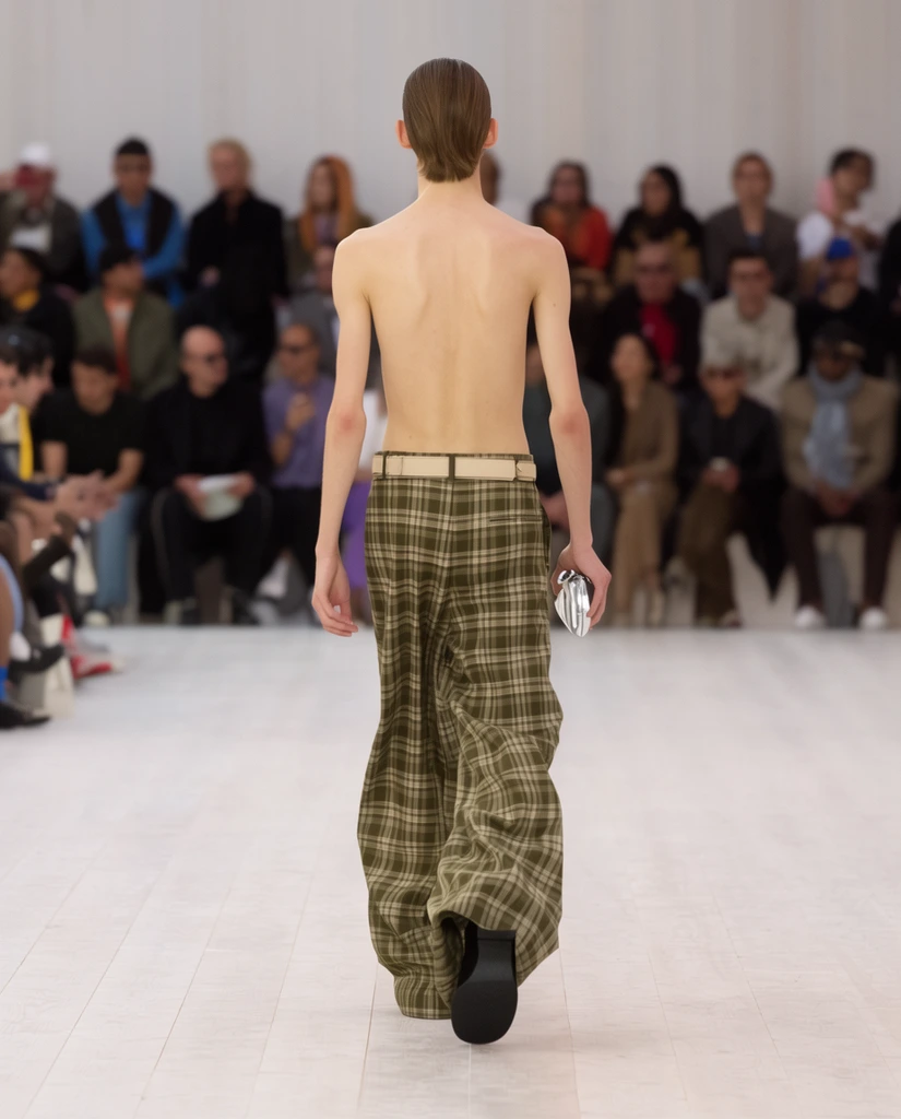 shirtless slender male model walking away on a fashion runway, extremely wide-leg olive green and beige plaid trousers, light beige belt, holding a small silver clutch, slicked-back brown hair, black shoe mid-stride, white wooden plank floor, blurred audience seated in background, shallow depth of field, telephoto lens, bright even lighting