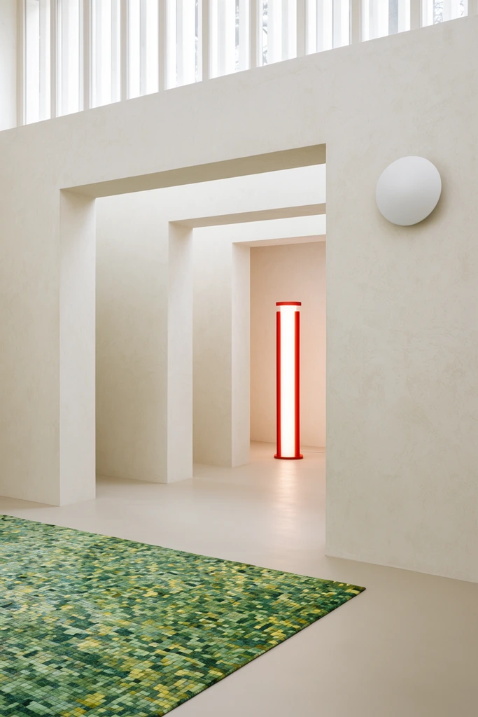 minimalist interior architecture, sequence of thick off-white plaster doorways creating deep perspective, soft natural daylight from high slatted windows, textured cream walls, bright red and white glowing cylindrical floor lamp in a recessed room, multi-tonal green and yellow mosaic patterned area rug, white circular wall sconce, smooth matte floors, architectural photography, serene gallery space, geometric composition, clean lines, high key monochromatic palette with vivid red and green accents