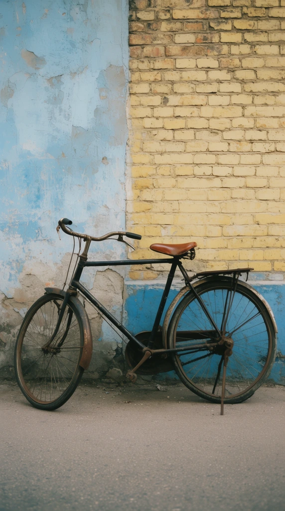 A vintage black bicycle with a brown leather seat rests on its kickstand against a dual-textured urban wall. The backdrop is divided vertically between crumbling light blue plaster on the left and faded yellow brick on the right. A lower band of blue paint grounds the brickwork. The bike features curved handlebars, rusted fenders, and a rear rack, standing on flat grey asphalt. The composition highlights weathered textures and rustic urban decay through natural daylight and a soft, medium-format film aesthetic.