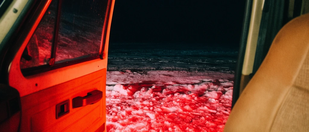 An open vehicle door frames a snowy night landscape illuminated by intense red light. From inside the cabin, a tan seat and an orange-tinted door panel establish a first-person perspective. Outside, rough snow glows with deep crimson hues, casting stark shadows across the icy texture. Beyond this brightly lit foreground, the flat terrain merges into a vast, pitch-black sky. The low-light photography features prominent film grain and extreme contrast between the warm artificial glow and the deep darkness.