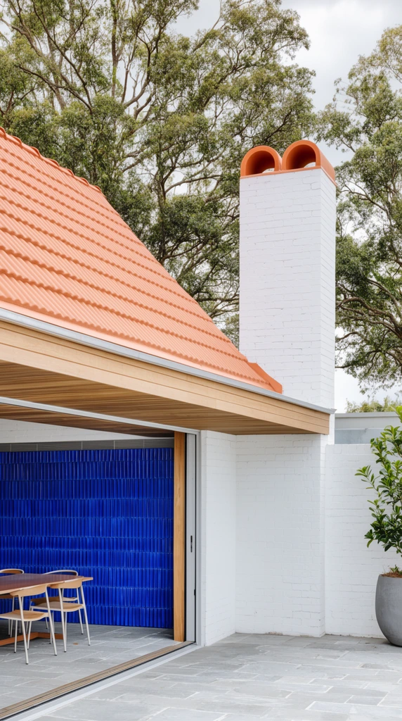 A modern outdoor patio framed by white painted brick walls and a steep terracotta ribbed roof with a wood-paneled overhang. Open sliding doors reveal a dining set against a striking vertical-ridged cobalt blue tiled wall. A towering white brick chimney with dual arched terracotta flues anchors the gray stone courtyard, backed by mature green trees under an overcast sky.