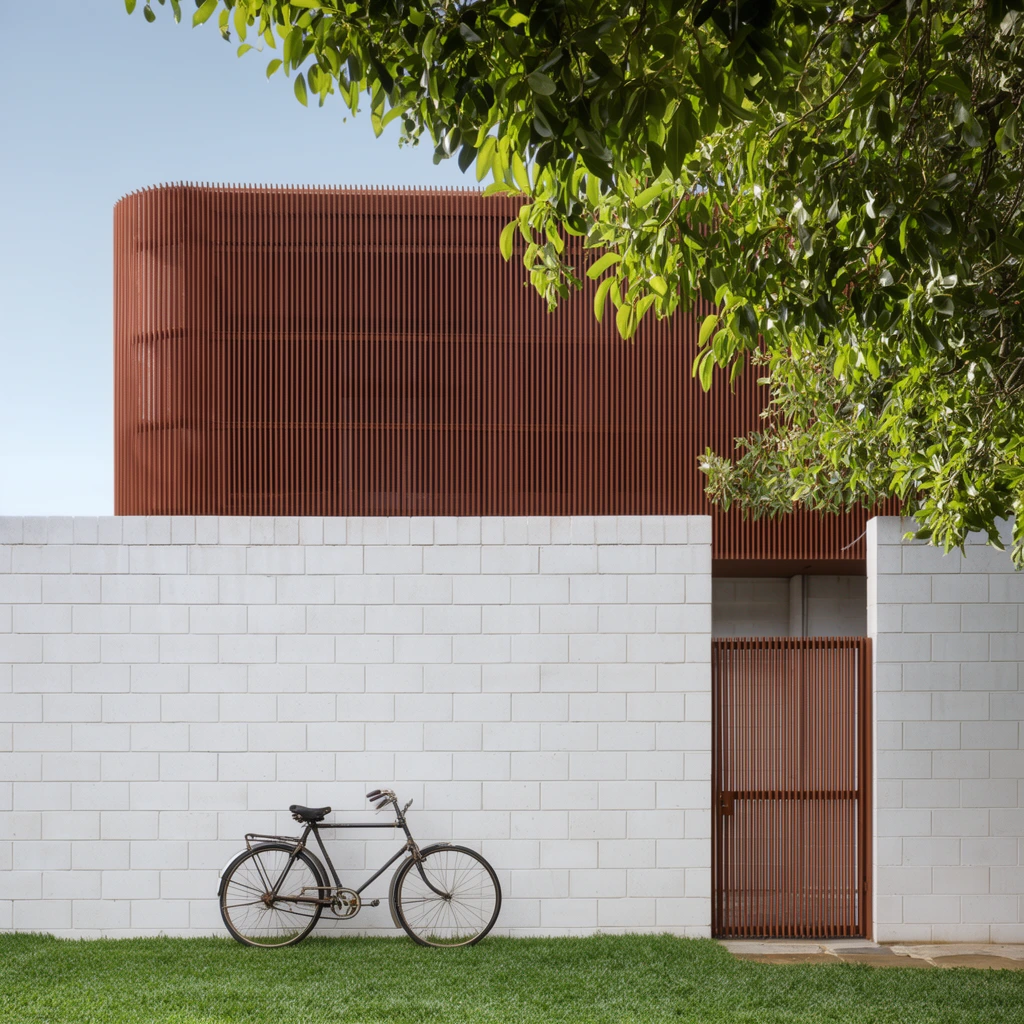 A modern two-story architectural exterior contrasting a white concrete block lower level with an upper volume wrapped in vertical rust-colored louvers. A dark vintage bicycle stands on bright green grass against the textured white wall. A matching slatted gate secures the ground floor to the right. The crisp composition is framed by lush green foliage overhanging from the top right against a clear pale blue sky. Architectural photography capturing natural daylight, sharp focus, and geometric minimalism.