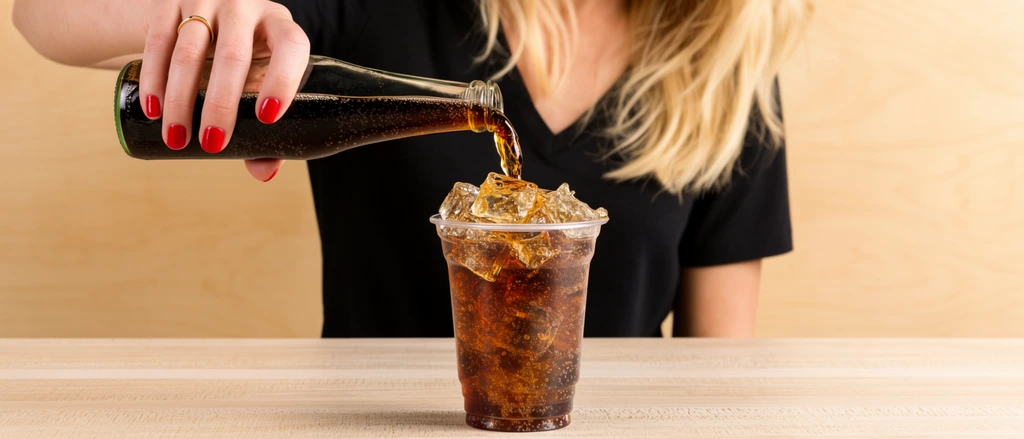close-up of a woman pouring dark fizzy liquid from a glass bottle into a clear plastic cup filled with ice, blonde hair, black v-neck shirt, bright red nail polish, thin gold ring, cup resting on a light wood textured table, smooth light wood paneled background, dynamic pouring action, liquid splashing over ice cubes, shallow depth of field, bright warm lighting, commercial food photography