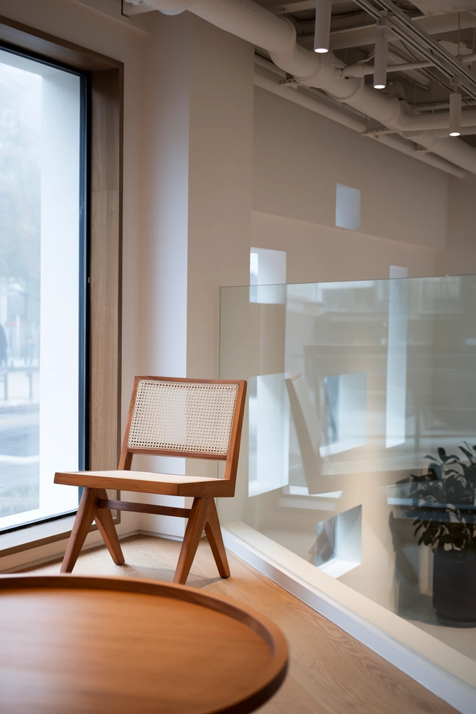 A minimalist interior space features a wooden cane chair positioned near a large window overlooking a multi-level architectural environment. In the foreground, the curved edge of a smooth round wooden table with a raised lip is slightly out of focus. Behind it, the chair rests on a light oak floor, distinguished by warm inverted V-shaped wooden legs and a tightly woven light rattan backrest. To the left, a tall window set in a wood frame reveals a hazy outdoor street scene. To the right, a transparent glass balustrade reflects the room and reveals a lower level featuring white walls, geometric architectural cutouts, and a dark potted plant. The ceiling exposes white-painted industrial pipes and track lighting with cylindrical fixtures. The architectural photography style utilizes soft, diffused natural daylight blending with warm overhead illumination, creating a serene color palette of crisp whites, warm woods, and soft grays with delicate glass reflections.