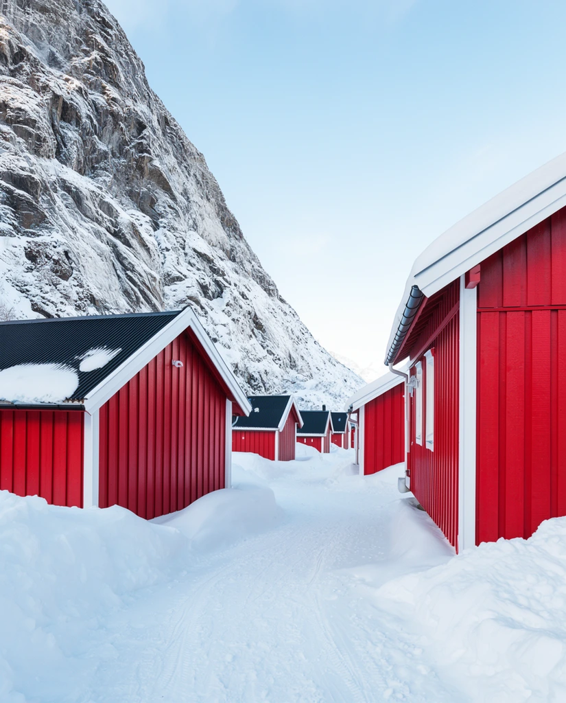 Vibrant red wooden cabins with white trim and snow-laden dark roofs line a thick snow-filled pathway. Deep pristine drifts bury the building foundations and reveal faint tracks. A towering, sheer rocky mountain face dusted with snow rises dramatically on the left, framed against a pale blue sky. Captured in crisp daylight with a wide-angle perspective, the serene landscape photography highlights the striking color contrast between the vivid crimson vertical paneling and the stark white winter environment.