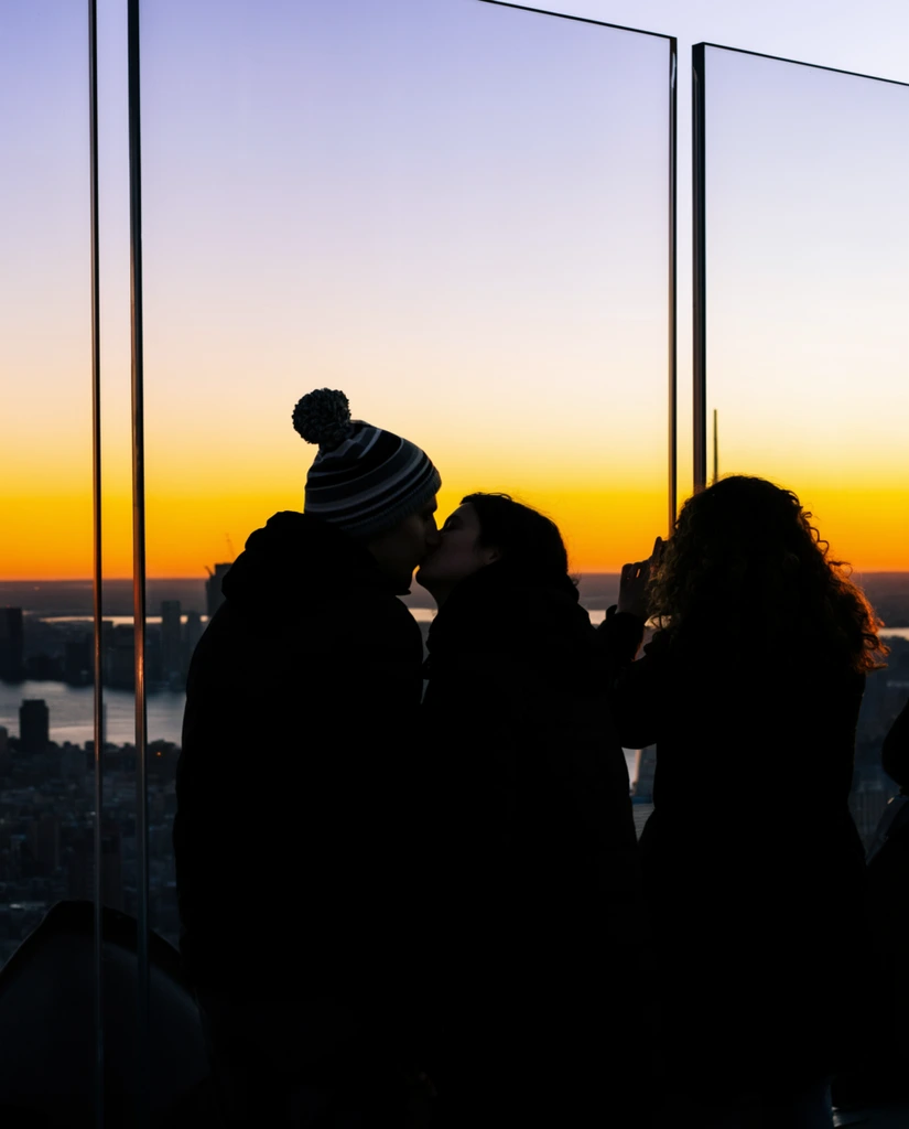 Two silhouetted figures share an intimate kiss on an elevated observation deck, framed against a brilliant sunset. The left figure wears a striped pom-pom beanie, both bundled in dark winter coats. Beside them, a third silhouetted person with long curly hair photographs the view. Tall transparent glass panels with stark vertical seams separate the dark foreground from a sprawling cityscape of skyscraper silhouettes and a winding river. The luminous sky forms a smooth gradient, transitioning from fiery golden-orange at the horizon to pale lavender and blue. High-contrast photography emphasizes the geometric lines and deep shadows against the vibrant, glowing background.