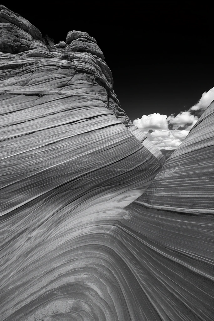 high-contrast black and white landscape of a sweeping sandstone formation, undulating sedimentary layers, continuous flowing parallel striations curving into a deep trough, smooth stone surfaces with dramatic shadows highlighting the sweeping contours, converging V-shaped rock walls, pitch-black sky with bright puffy clouds near the right horizon, wide-angle perspective, sharp focus monochrome