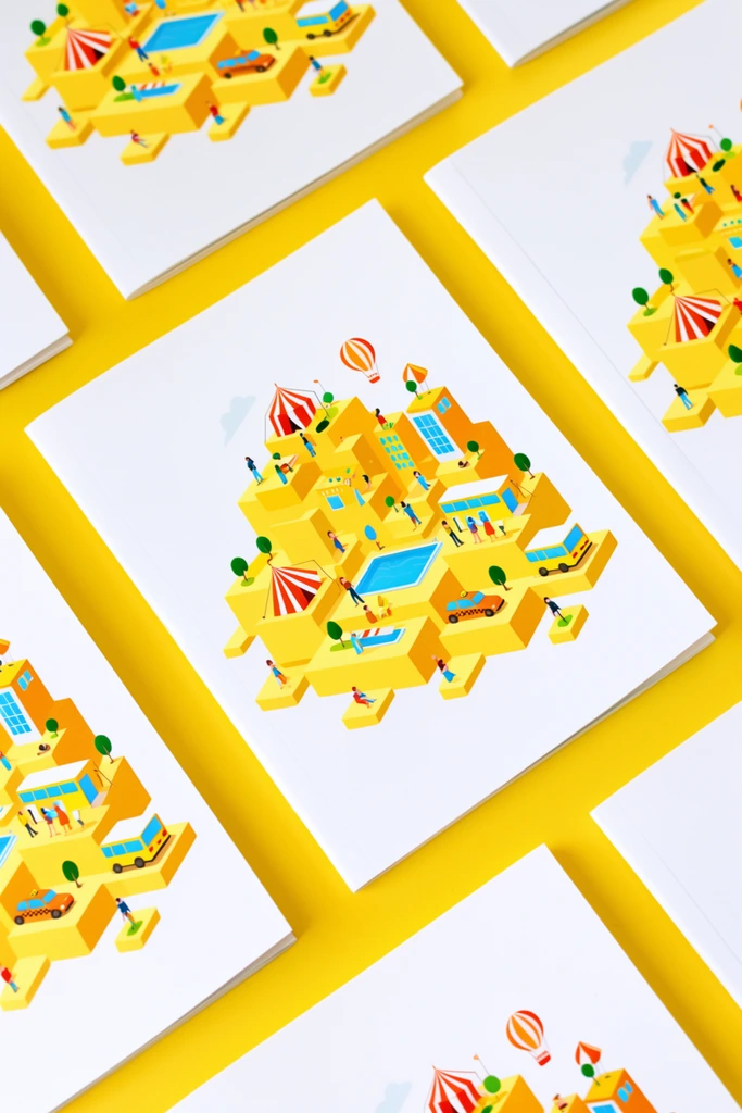 Multiple white printed booklets rest diagonally on a vivid yellow surface. The covers feature a colorful isometric vector illustration of a miniature cityscape built on dimensional yellow geometric blocks. Tiny stylized figures populate the scene alongside a blue swimming pool, a yellow bus, an orange taxi, striped tents, small trees, and a hot air balloon. The modern design utilizes crisp lines and a bright palette of cyan, orange, and red. The composition is captured from a top-down, slightly angled perspective with even lighting.