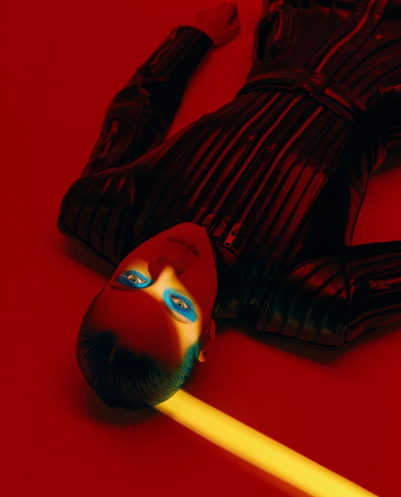 A top-down view of a person lying on a vibrant crimson surface wearing a structured, ribbed black leather garment with metallic zippers. The subject features slicked-back dark hair with a teal sheen and striking cyan-blue eye makeup. The entire scene is heavily saturated in deep red ambient lighting. A stark diagonal beam of intense golden-yellow light cuts sharply across the lower frame, illuminating a precise band across the subject's face and highlighting the vibrant blue makeup. This high-contrast, directional lighting contrasts the glossy dark leather against the uniform red background, creating a bold, cinematic fashion editorial aesthetic defined by intense color blocking and sharp geometric shadows.