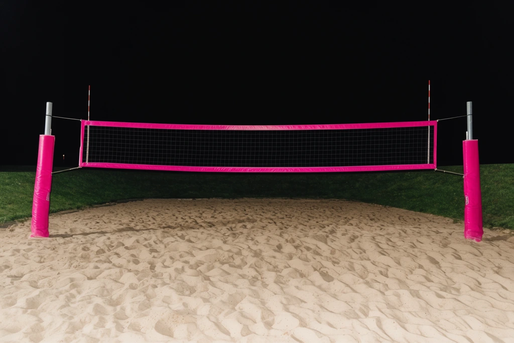 Centered sand volleyball net at night, vibrant pink borders and padded poles, harsh flash lighting, textured sand foreground, dark grassy berm, pitch black sky, stark high-contrast photography