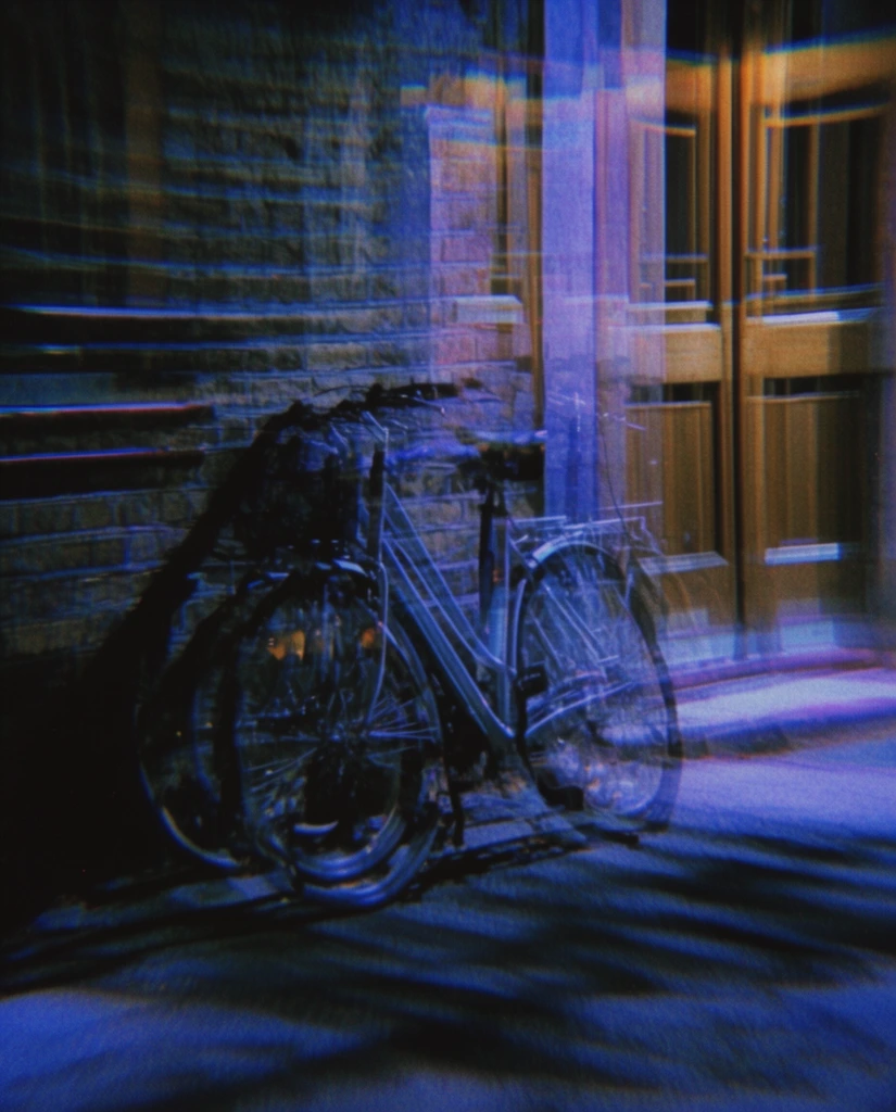 A bicycle leaning against a brick wall on a paved street next to a double wooden door is rendered with a heavy, horizontal multiple-exposure and severe motion blur effect. The bicycle's structure, the textured brick wall, and the vertical panels of the double wooden door situated to the right appear duplicated in multiple semi-transparent overlapping layers stretching laterally across the frame. The paved street features dark, streaking shadow patterns. The nocturnal scene utilizes a highly saturated color palette dominated by deep indigo blues, vibrant electric purples, and warm tungsten-colored highlights glowing against stark, crushed black shadows. The photographic technique applies a kaleidoscopic prism distortion, resulting in ghostly, phantom-like repetitions of the scene's geometry and pronounced horizontal light trails. The overall aesthetic mimics high-ISO film photography, exhibiting heavy coarse grain, color noise, chromatic aberration, and a moody, disorienting atmosphere characteristic of experimental 90s nocturnal cinematography.