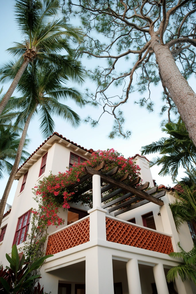 A low-angle view captures a white stucco Mediterranean-style building framed by towering palm trees and sprawling branches. The architectural structure features a red terracotta roof, dark red window frames, and a prominent second-story terrace enclosed by a decorative terracotta lattice balustrade. A wooden pergola spanning the terrace is heavily draped with vibrant pink bougainvillea vines. Lush green palm fronds and a massive, textured tree trunk on the right arch upward across a clear pale blue sky. Soft natural daylight highlights the crisp facade and rich tropical foliage.