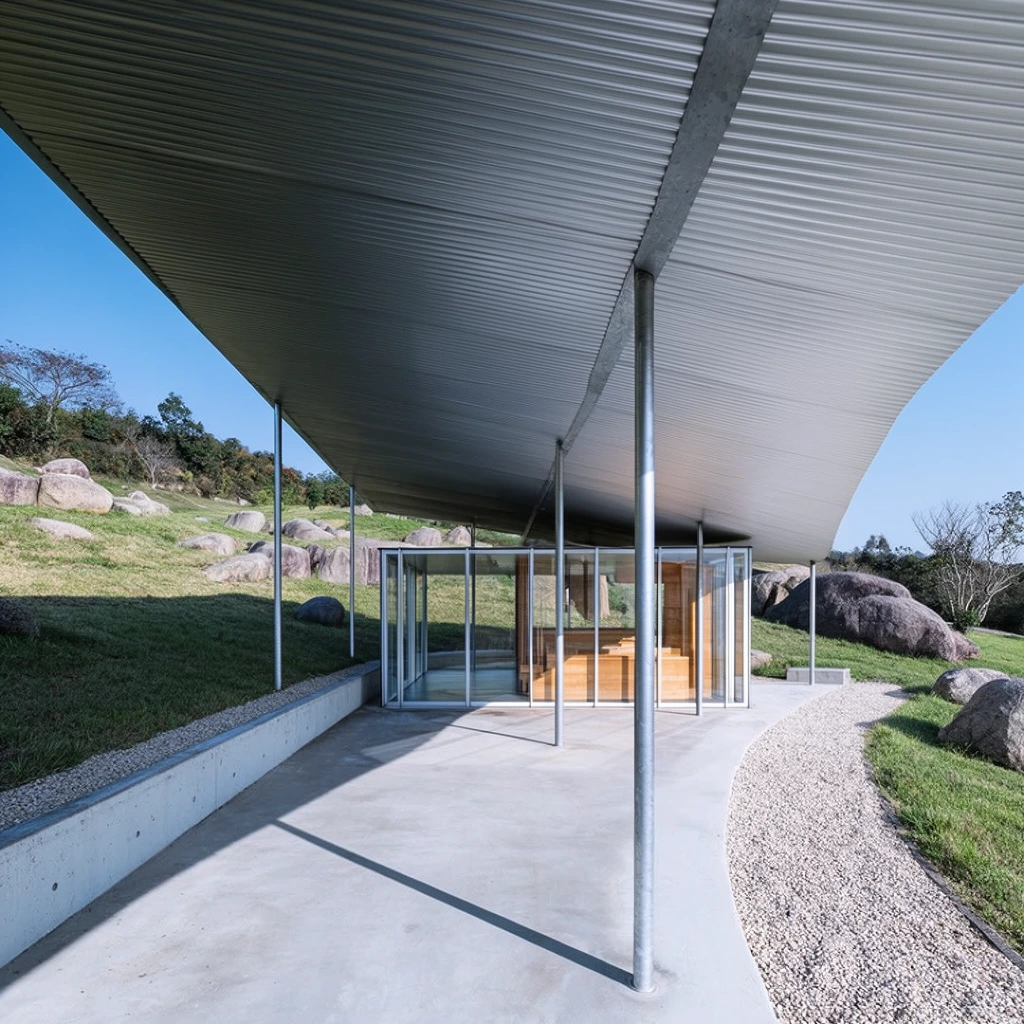 A minimalist architectural pavilion featuring a wide corrugated metal canopy supported by slender cylindrical steel columns. Beneath the expansive roof, an enclosed room with floor-to-ceiling glass walls reveals warm wooden interior elements. The structure rests on a smooth concrete patio bordered by a low concrete wall and a curved light gravel walkway. Sharp, high-contrast geometric shadows stretch across the concrete floor under bright direct sunlight. The pavilion sits on a sloping hillside covered in green grass, dotted with large rounded grey boulders and distant trees against a clear blue sky. The architectural photography employs striking linear perspective, drawing the eye along the shaded concrete edge toward the illuminated glass enclosure.