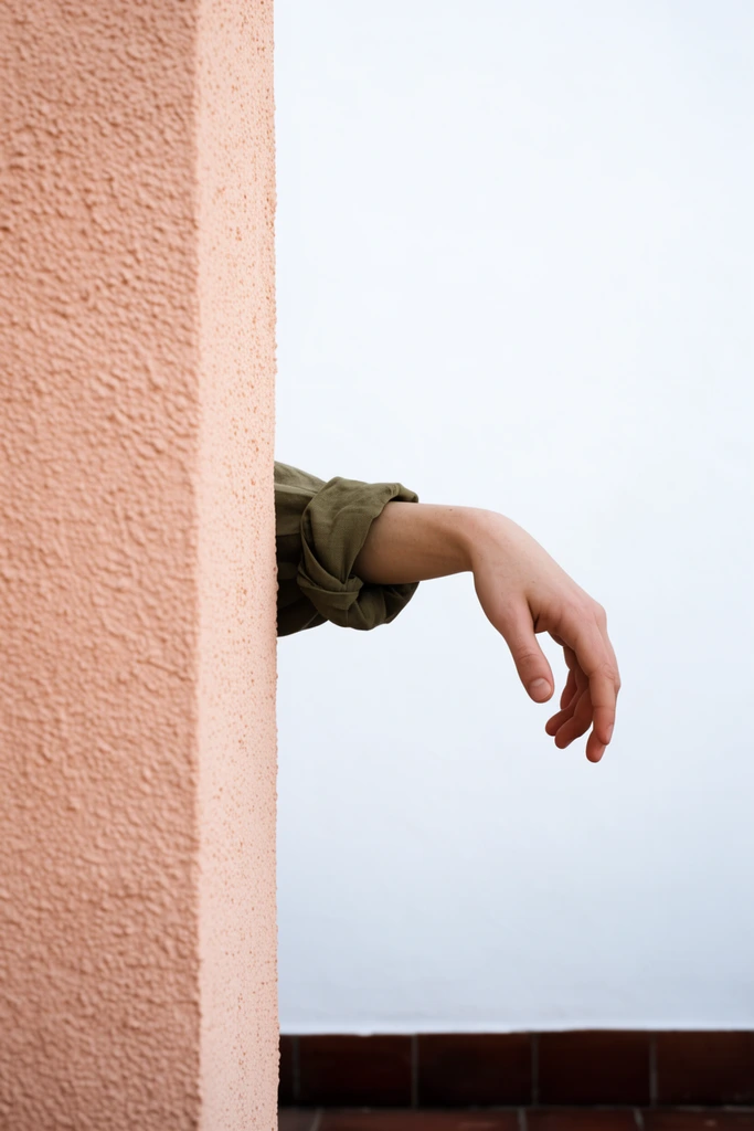 A solitary arm with a rolled olive sleeve extends horizontally from behind a highly textured peach wall. The relaxed hand hovers against a stark white background above dark terracotta floor tiles, forming a surreal minimalist composition.