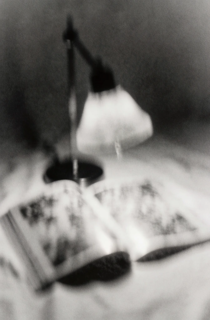 ethereal monochromatic still life, heavy film grain, extreme soft focus, blurred desk lamp suspended over an open book, ghostly textures, high contrast lo-fi aesthetic, mottled surface noise, vintage black and white palette, dreamlike impressionistic atmosphere, hazy outlines, nostalgic and haunting mood, minimalist composition, textured paper and lamp finish obscured by intentional blur, soft atmospheric lighting, grainy film photography style.