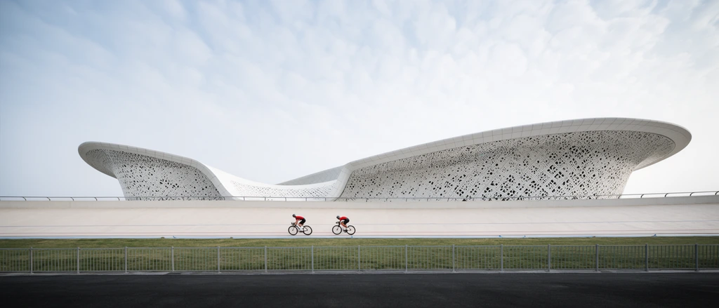 two cyclists in red and black gear riding on a light concrete banked track, sweeping modern white architectural building with undulating curved roof, intricate perforated geometric facade, dark asphalt foreground separated by a metal fence, grassy berm, pale cloudy sky, soft daylight, wide architectural sports photography