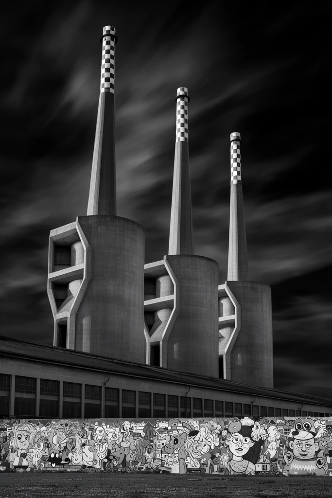 three towering brutalist concrete smokestacks, cylindrical bases featuring large angular cutouts, tapered spires with checkered caps, long low industrial building below, foreground wall densely covered in illustrative cartoonish street art, dark dramatic sky with sweeping motion-blurred clouds, high contrast black and white architectural photography, long exposure