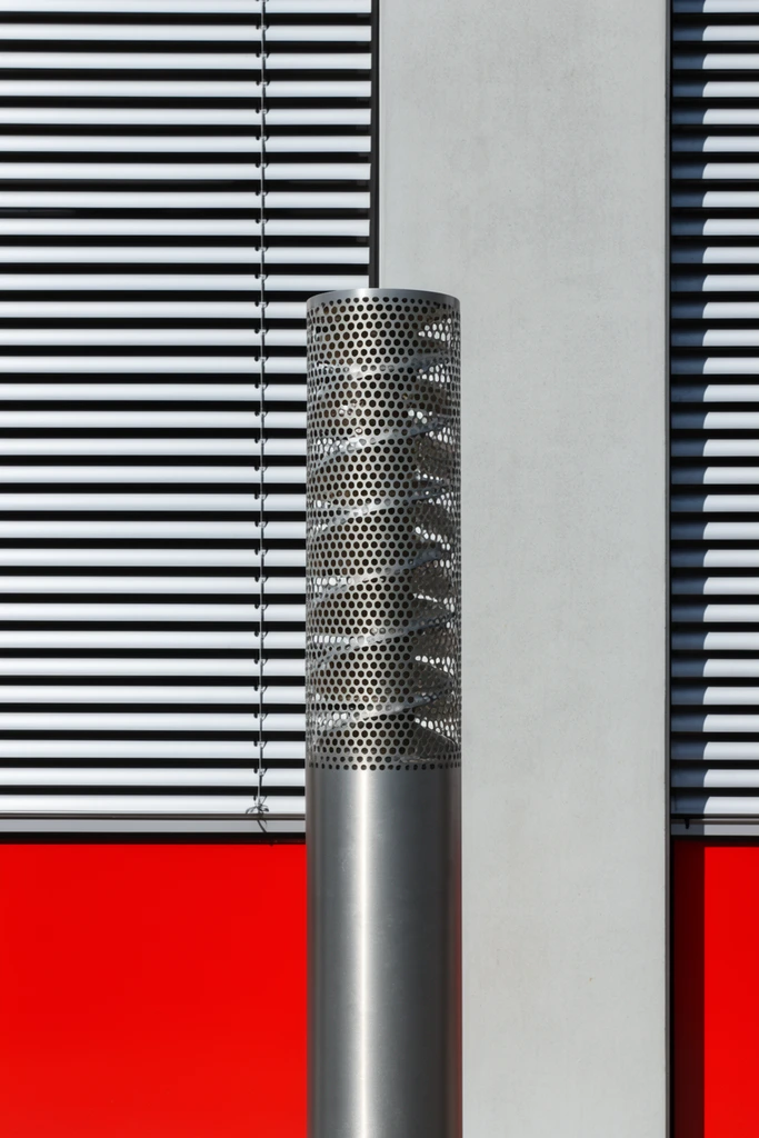 cylindrical metallic bollard featuring a perforated upper section with an internal spiral structure, set against a minimalist architectural facade, horizontal silver window blinds forming high-contrast black shadow stripes, flat light gray concrete vertical pillar, vibrant solid red horizontal band spanning the lower third, geometric abstraction, hard daylight, sharp focus, interplay of intersecting perpendicular lines
