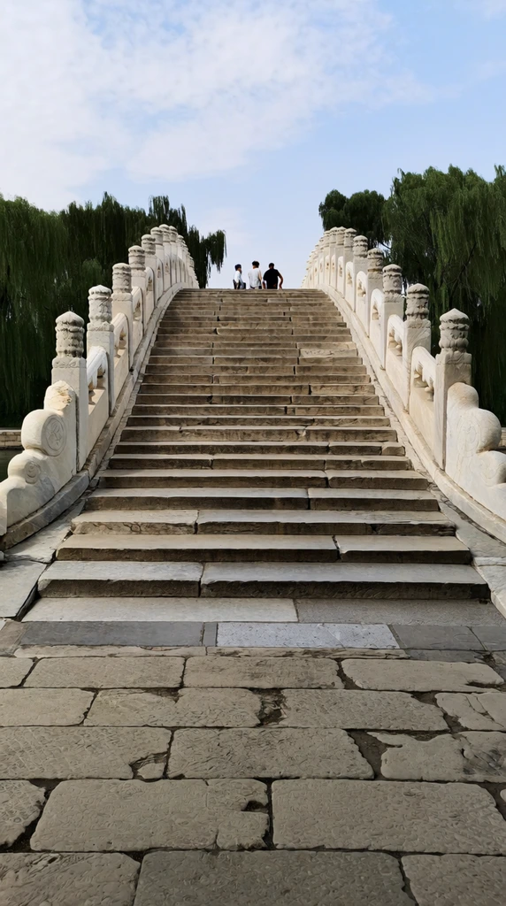 steep white marble arch bridge, ornate balustrades with carved stone pillars, weathered grey stone steps ascending a sharp incline, three distant figures standing at the crest, large textured paving slabs in foreground, lush weeping willow trees, pale blue sky with scattered clouds, natural daytime lighting, architectural landscape photography