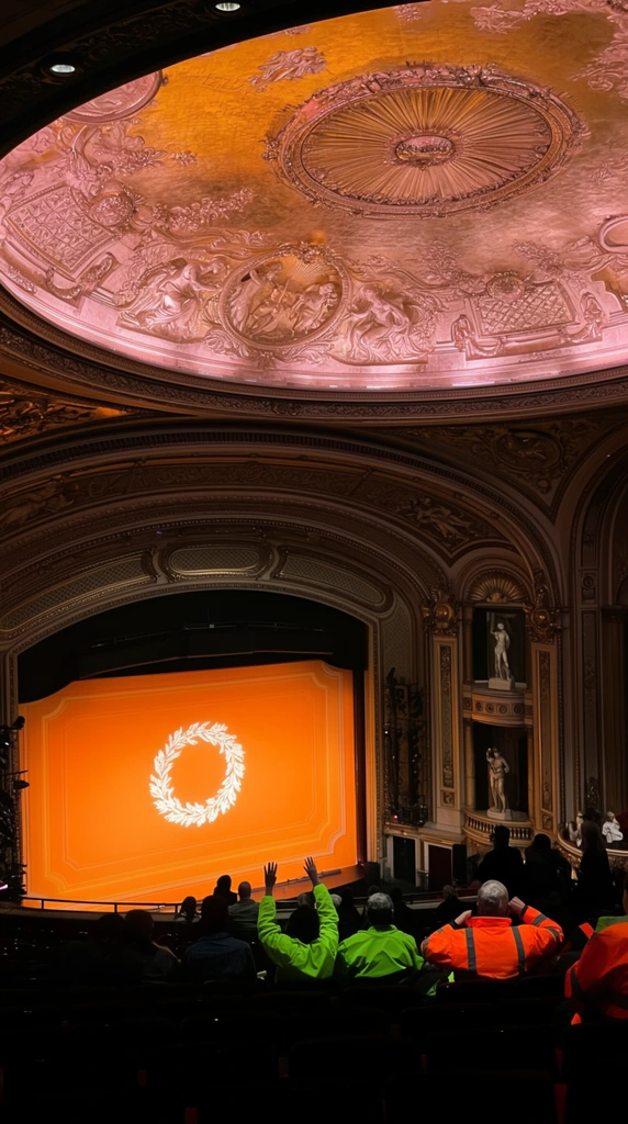 A grand theater interior viewed from a high balcony showcases a magnificent gold-leaf dome ceiling and a brightly illuminated stage. The massive recessed circular dome is ornately decorated with classical bas-relief sculptures, warm rosy lighting, and a central sunburst fixture. Below sweeping arched moldings, the proscenium frames a vibrant orange stage backdrop centered with a glowing white wreath motif. In the darkened lower foreground, silhouetted audience members sit in rows, punctuated by individuals wearing bright neon green and high-visibility orange jackets, one raising both hands. To the right, tall side boxes house classical statues. The wide-angle photograph contrasts deep auditorium shadows with luminous gold and amber tones highlighting the intricate architecture and intense stage lighting.