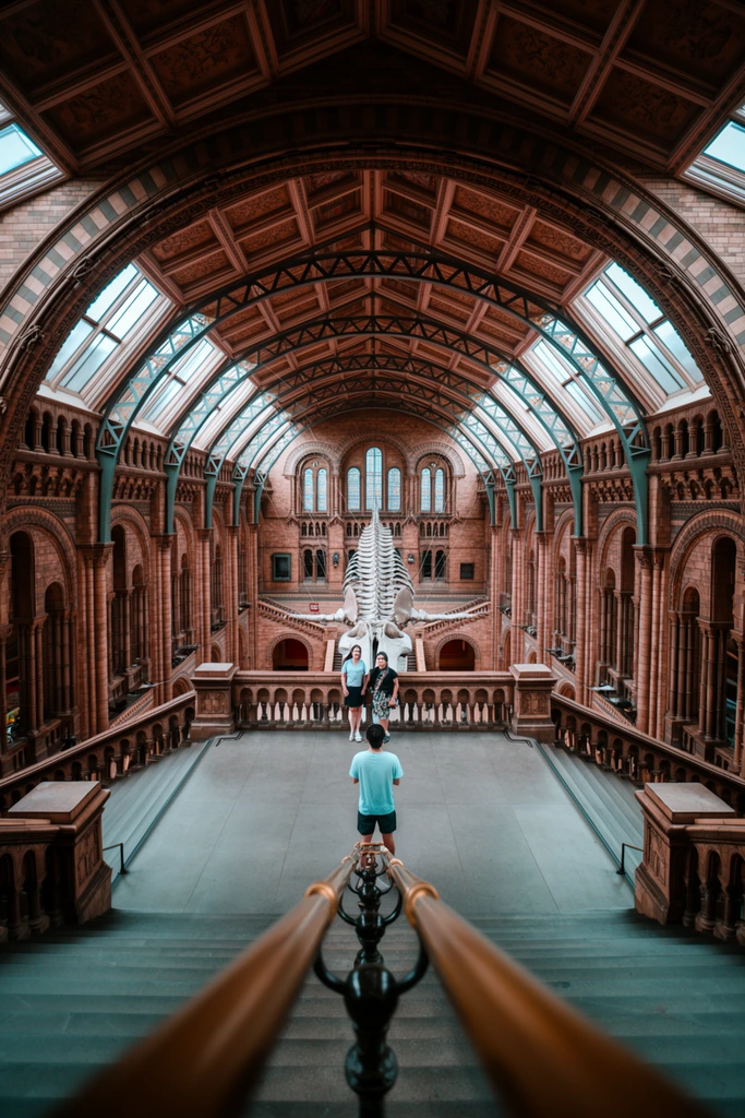 A photographer captures a portrait of two companions on a central balcony overlooking a monumental Victorian-era museum hall. Blurred brass handrails in the extreme foreground lead down a grand stone staircase toward the photographer, clad in a light blue t-shirt and dark shorts. Beyond the ornate stone balustrade, a colossal whale skeleton hangs suspended in the cavernous, perfectly symmetrical space. The soaring architecture features intricate terracotta brickwork, majestic rounded archways, exposed green iron trusses, and expansive glass skylights filtering soft daylight into the grand interior. Rich amber, warm brown, and muted teal tones dominate the wide-angle architectural composition, emphasizing the sweeping scale of the arched ceiling and the dramatic contrast between the massive skeletal display and the small human figures.