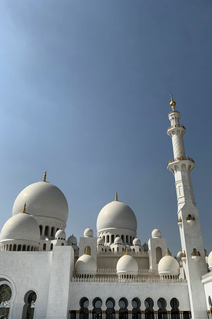 A grand white marble mosque features smooth, clustered domes and a towering minaret against a clear blue sky. Gold finials crown the domes. Bright daylight highlights arched windows and tiered balconies from a low angle.