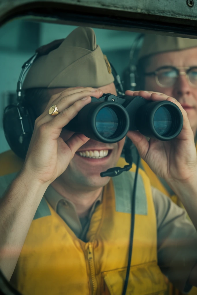 smiling man looking through large black binoculars, right hand holding optics revealing a gold signet ring on pinky, broad teeth-baring smile, tan garrison cap, bright yellow life vest, dark headphones, throat microphone, out-of-focus background figure in matching cap and wire-rimmed glasses, framed by dark structural window, subtle glass reflections, cinematic mid-shot, shallow depth of field, vintage color grading, teal and warm tones