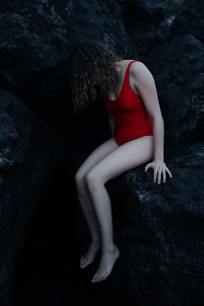 woman with wet curly hair obscuring her face, sitting precariously on massive dark jagged volcanic rocks, vivid red one-piece swimsuit contrasting sharply against pale skin and deep black shadows, bare legs dangling into a pitch-black crevice, hand gripping the rock edge, low-key lighting, moody atmospheric photography, dramatic chiaroscuro, cool tones, highly textured stone surfaces