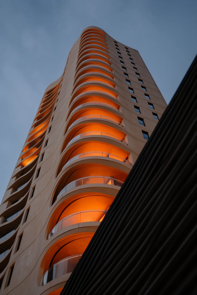 A low-angle perspective looking up at a towering modern high-rise against a muted blue dusk sky. The skyscraper features an undulating facade of smooth beige concrete. Recessed, curved balconies stack vertically along the structure, each illuminated from within by an intense, warm orange glow and enclosed by sleek glass railings. Small dark windows punctuate the flat concrete sections. In the lower right foreground, a dark, deeply corrugated architectural element cuts diagonally across the frame, creating stark geometric contrast to the sweeping curves of the tower. The composition highlights a dramatic interplay between cool ambient twilight and vibrant artificial warmth.